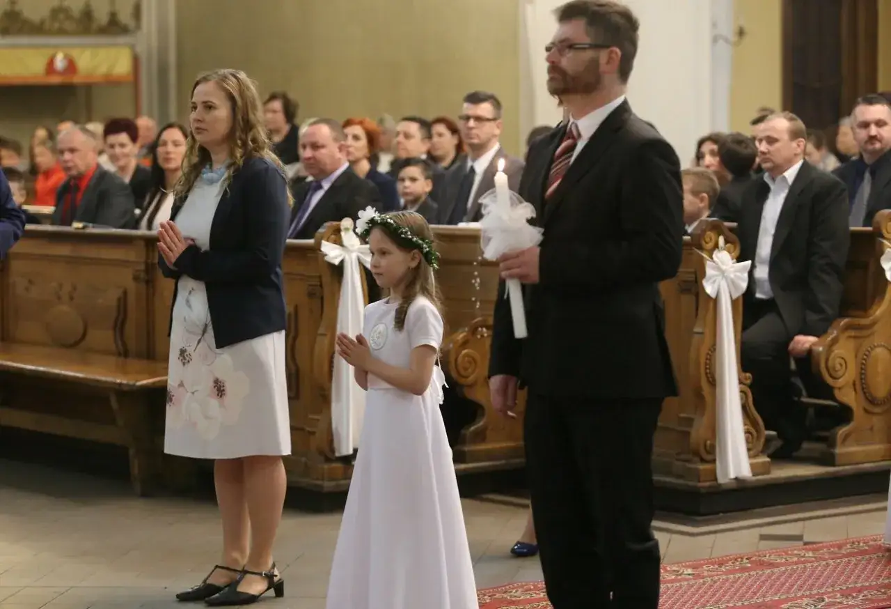 Elegancka sukienka w kwiaty i granatowa marynarka to dobry pomysł, jak się ubrać na komunię. Dziewczynka w bieli z wiankiem na głowie i mężczyzna z zapaloną świecą.