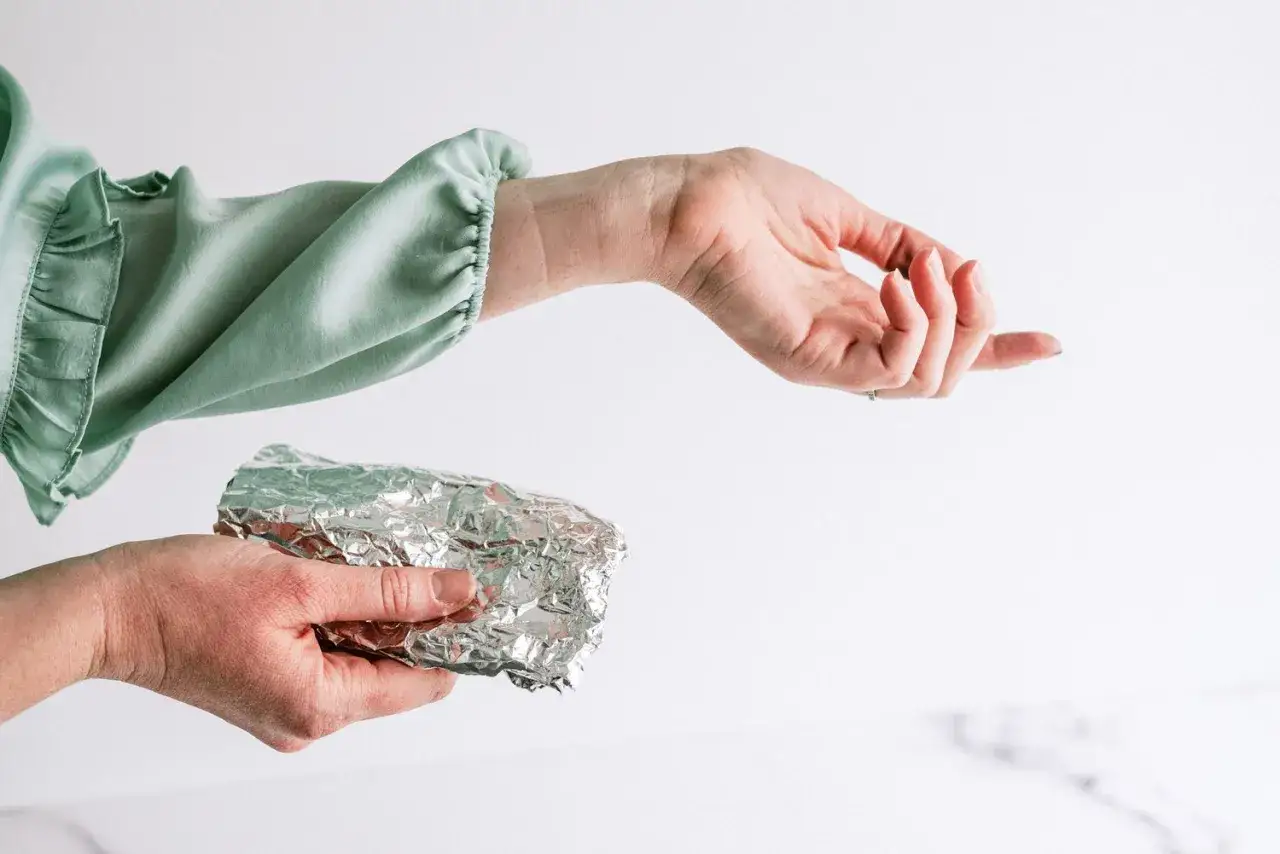 Eine Hand h&auml;lt ein St&uuml;ck Alufolie, w&auml;hrend eine andere Hand mit einem mintgr&uuml;nen, gerafften &Auml;rmel daneben schwebt. Vielleicht ein Trick gegen statisch aufgeladene Kleidung?