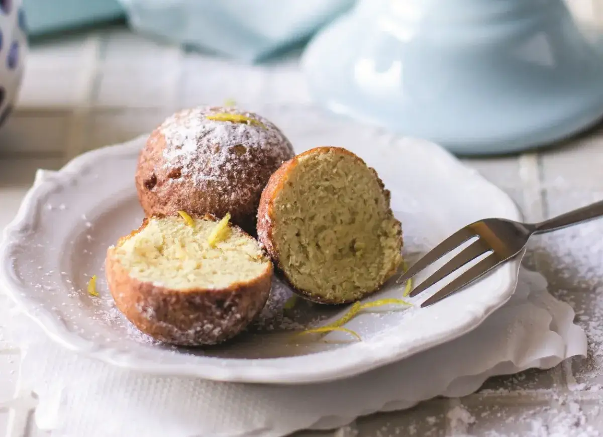 Puszyste paczki z serka homo, posypane cukrem pudrem i skórką cytrynową, kuszą swoim domowym smakiem. Jedna przekrojona ukazuje wilgotne wnętrze.