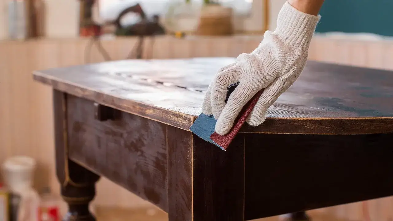 Mano guantata leviga il bordo di un tavolo scuro, preparando la superficie per la cementite senza carteggiare.