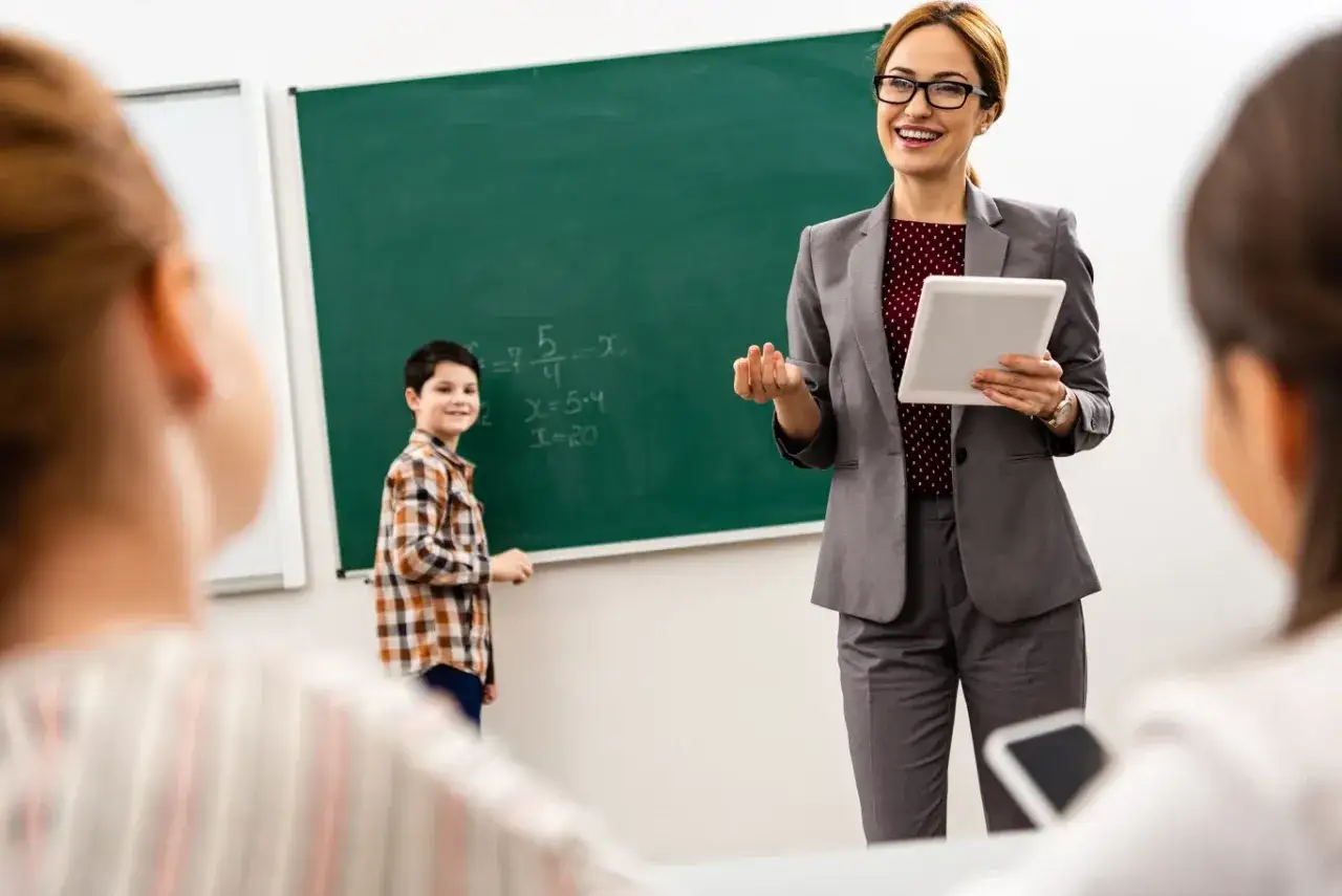 Uśmiechnięta nauczycielka z tabletem prowadzi lekcję matematyki. Uczeń rozwiązuje zadanie przy tablicy. Nawet przy niskich zarobkach nauczycieli, ich praca jest ważna.