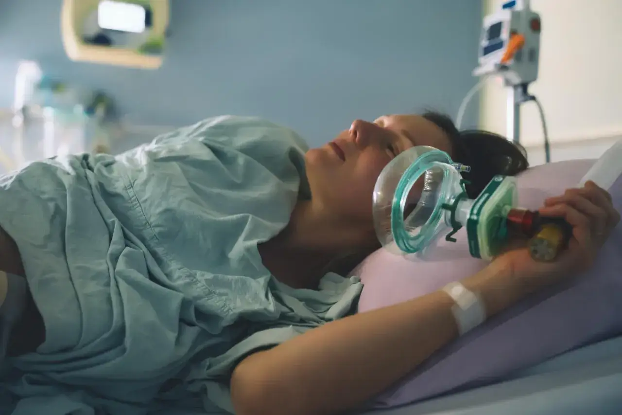 Kobieta w szpitalnym ł&oacute;żku, trzymająca maskę z gazem rozweselającym podczas porodu.