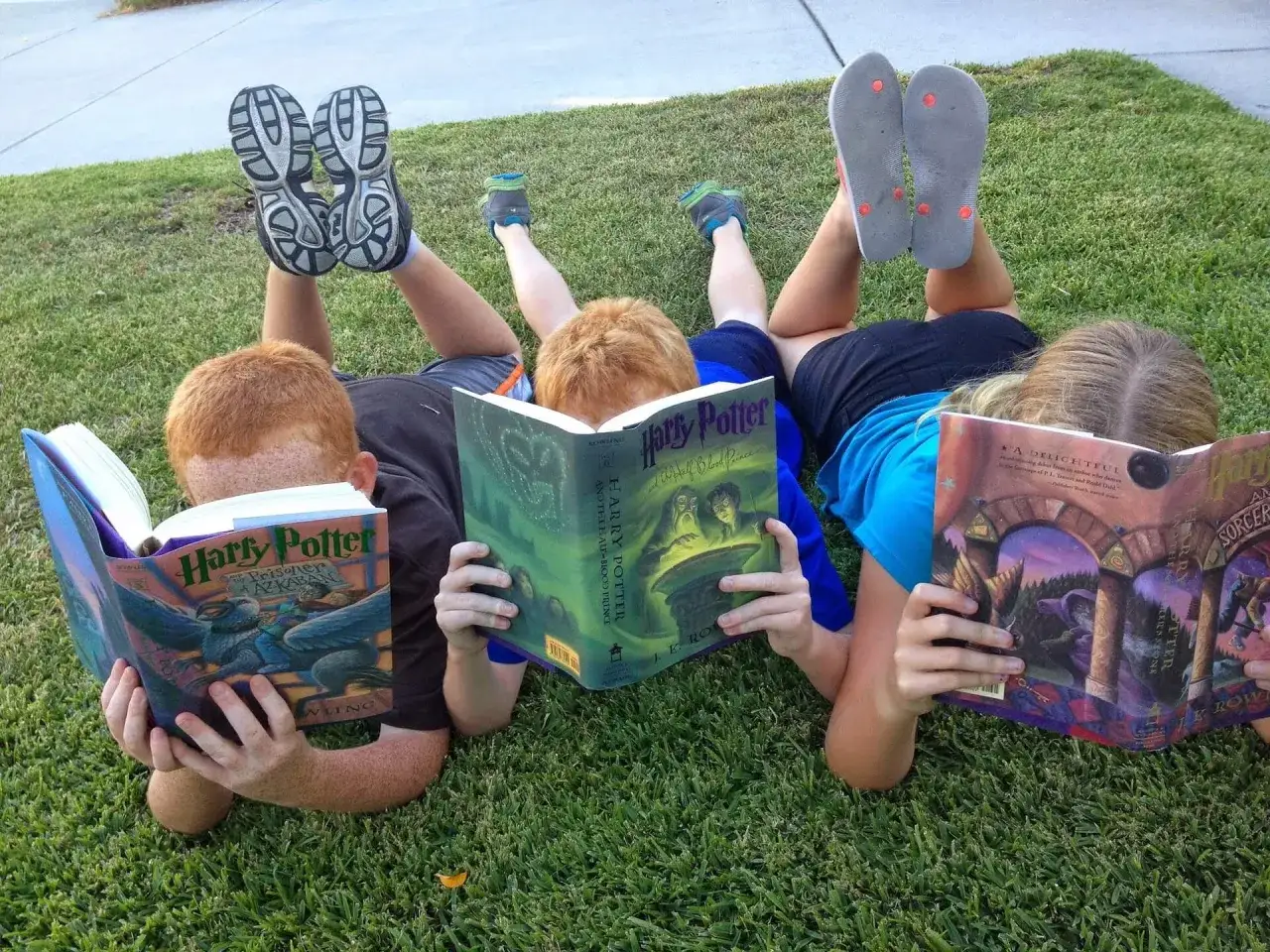 Trójka dzieci leży na trawie, czytając książki o Harrym Potterze. Ich nogi wyciągnięte są w górę, jakby sięgały drzewa do samego nieba.