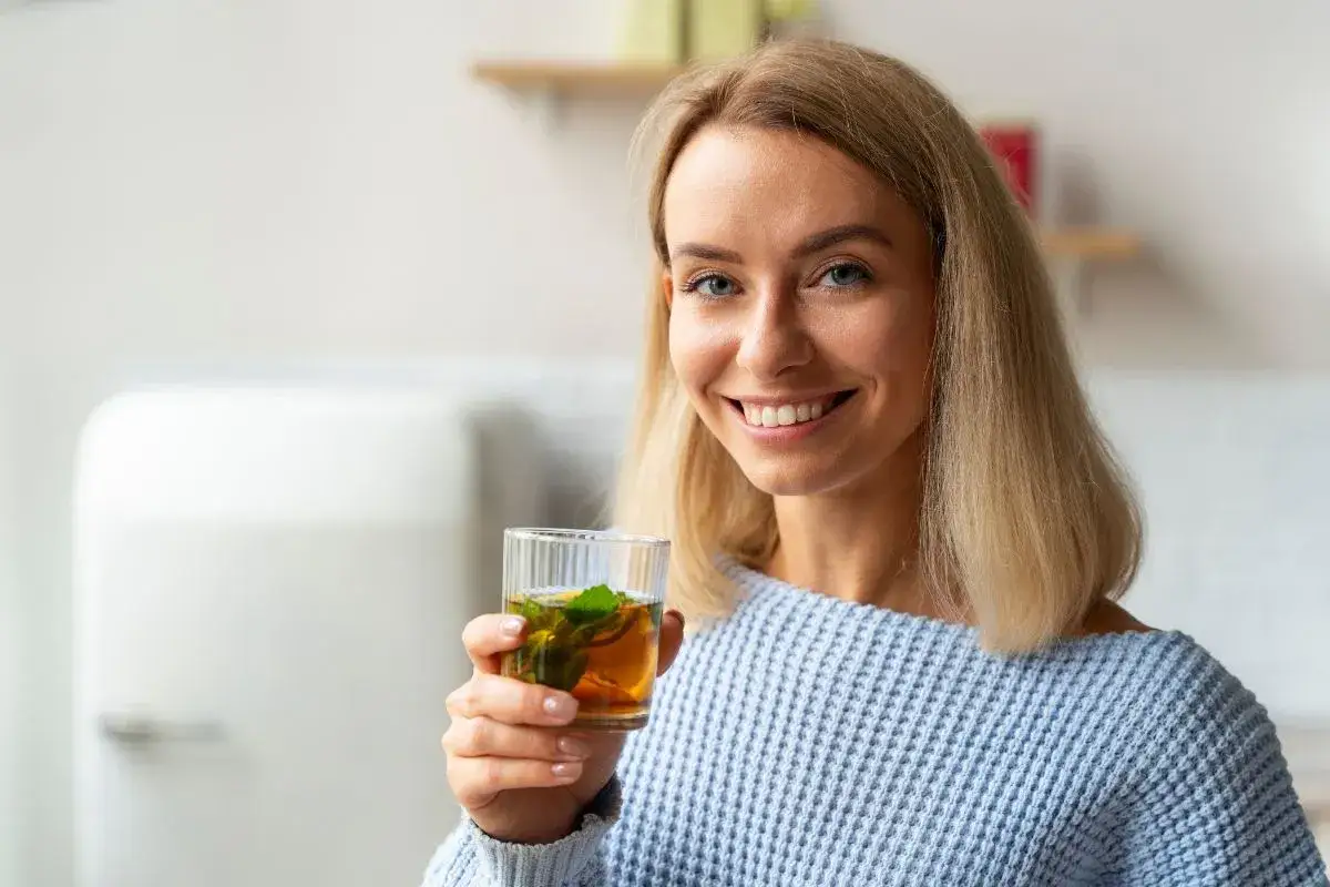 Uśmiechnięta blondynka trzyma szklankę z herbatą miętową. Czy herbata odwadnia? To pytanie zadaje sobie wiele osób, pijąc ten orzeźwiający napój.