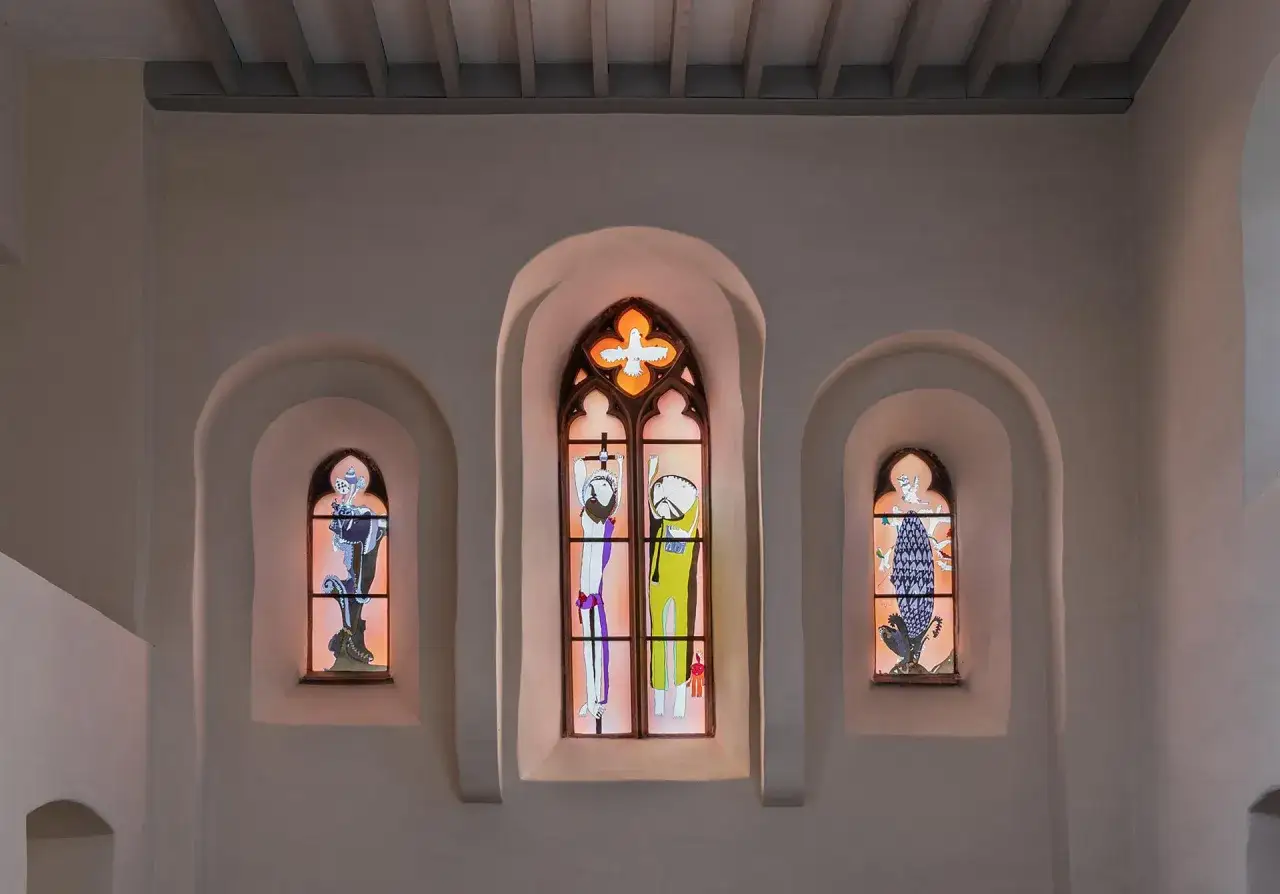 Drei bunte Glasfenster in einer Kirche. Das mittlere Fenster zeigt zwei Figuren, die an Kreuze gebunden sind, und dar&uuml;ber eine Taube, die das Gewissen symbolisiert.