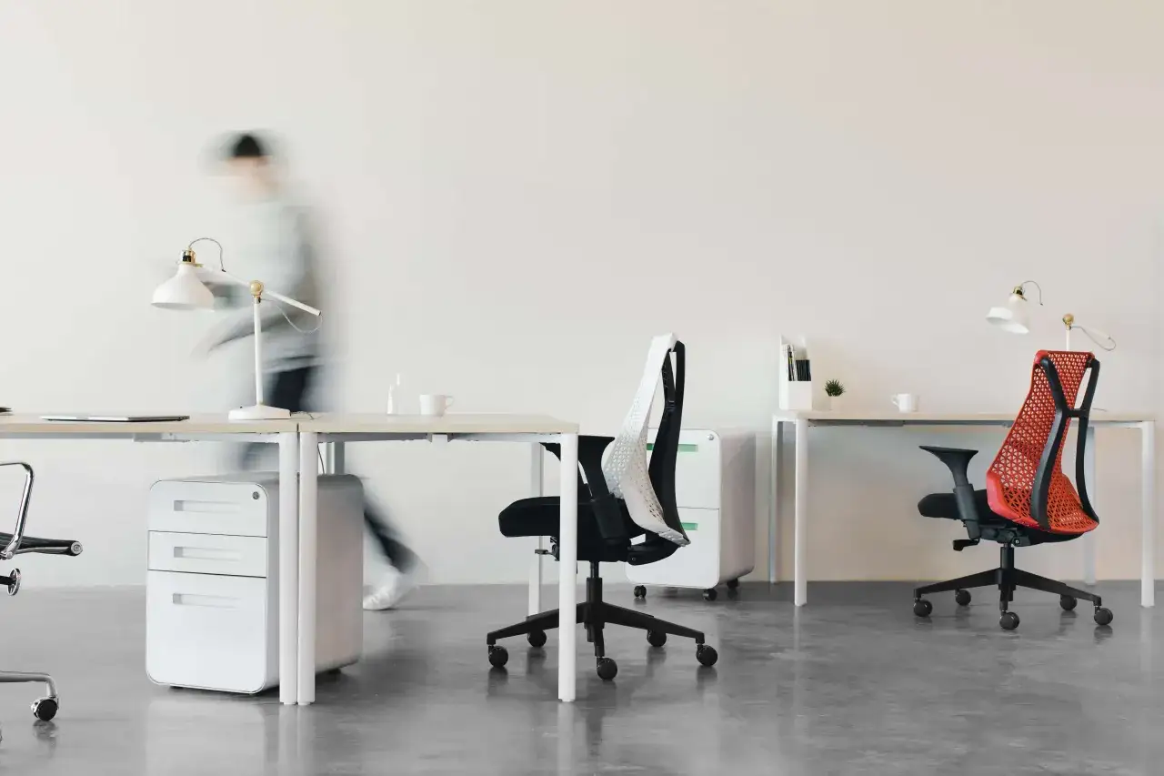 Espace de travail moderne avec deux bureaux dos &agrave; dos. Une personne floue passe, des chaises de bureau et des lampes sont visibles.