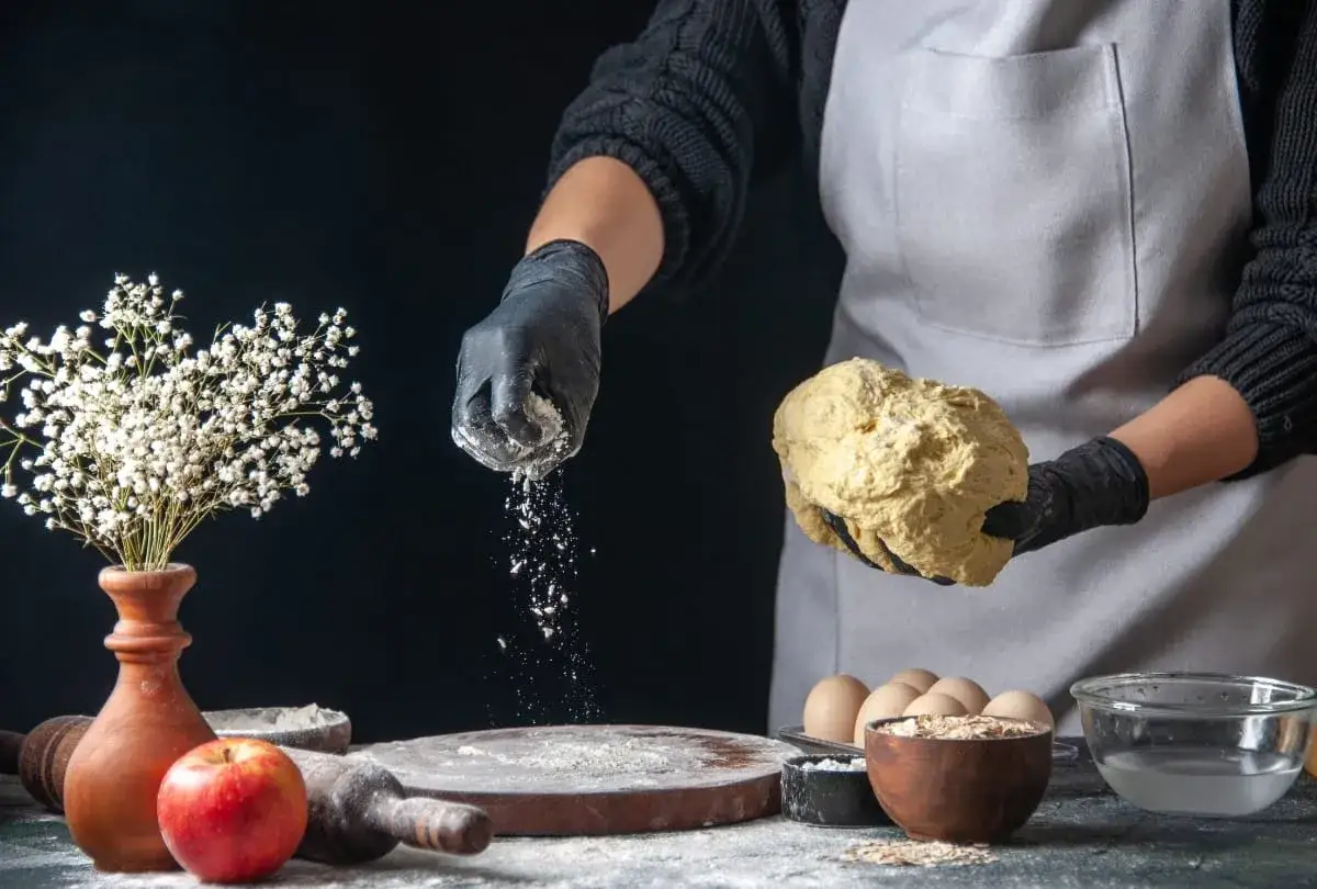 Hände mit Handschuhen streuen selbsttreibendes Mehl über Teig. Ein Apfel und Blumen sind auf dem Tisch.