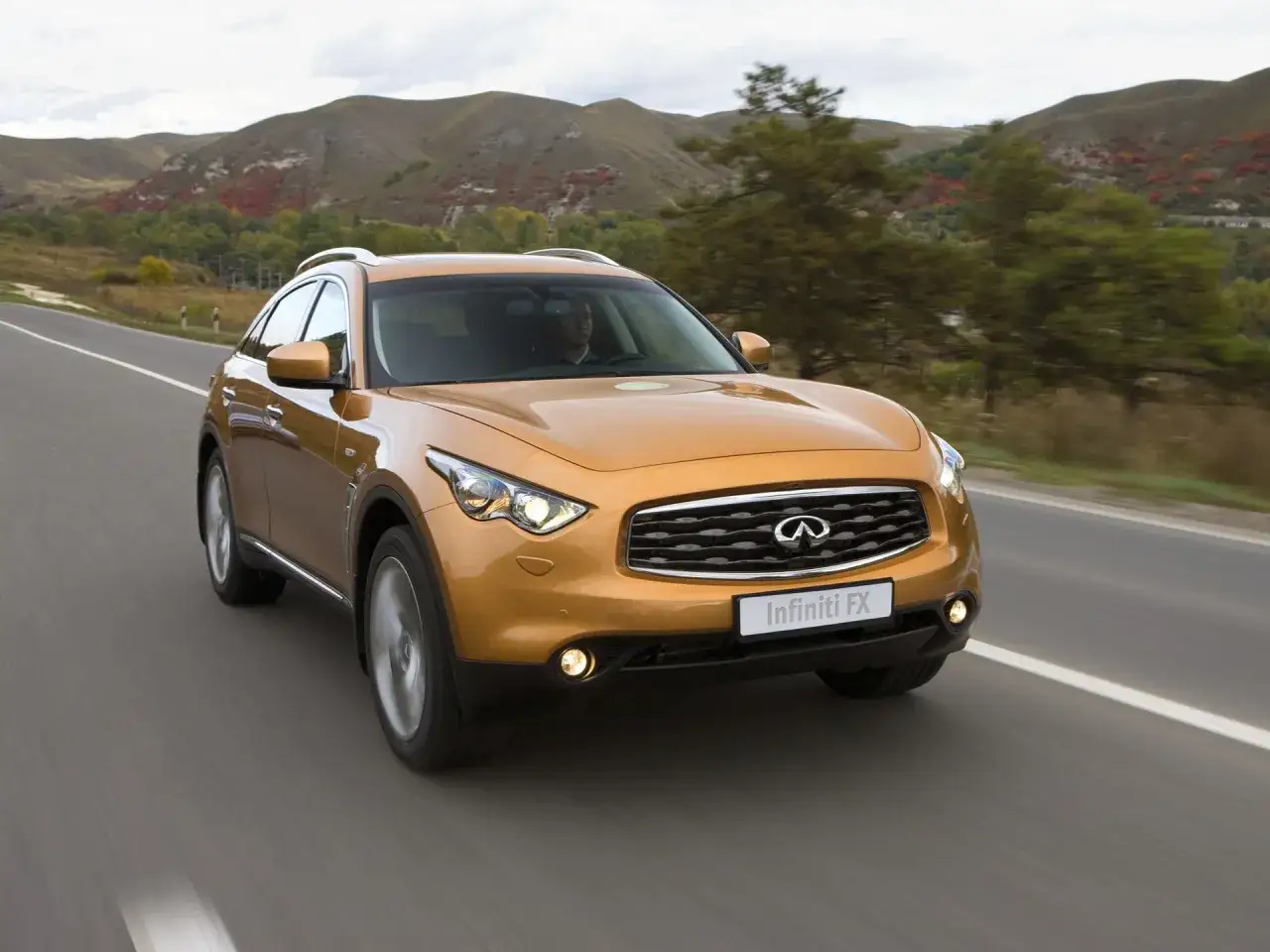 Pomarańczowy Infiniti QX70 jedzie po krętej drodze wśród jesiennych wzgórz.