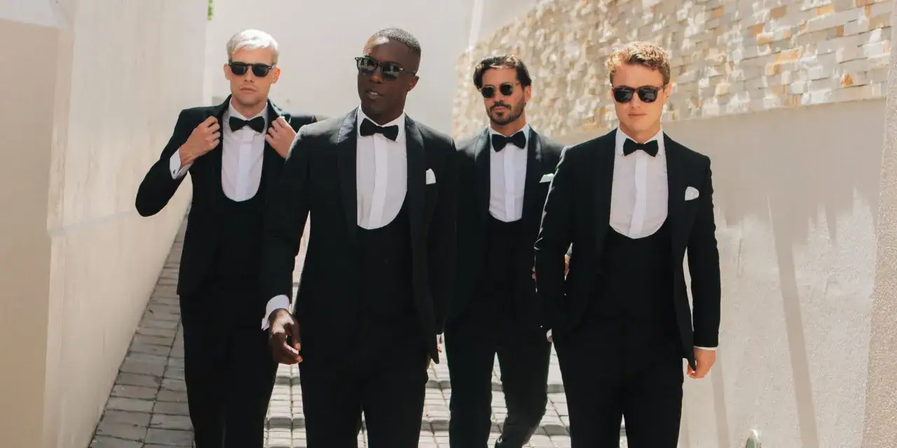 Four men in sharp tuxedos with cummerbunds walk together, looking dapper and ready for a night out.