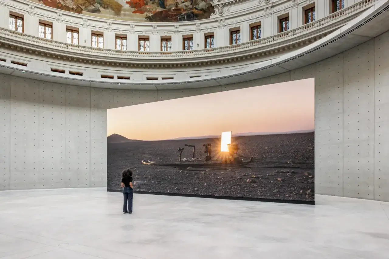Une femme contemple une œuvre d'art monumentale dans l'expo Pinault, représentant un paysage désertique au coucher du soleil.