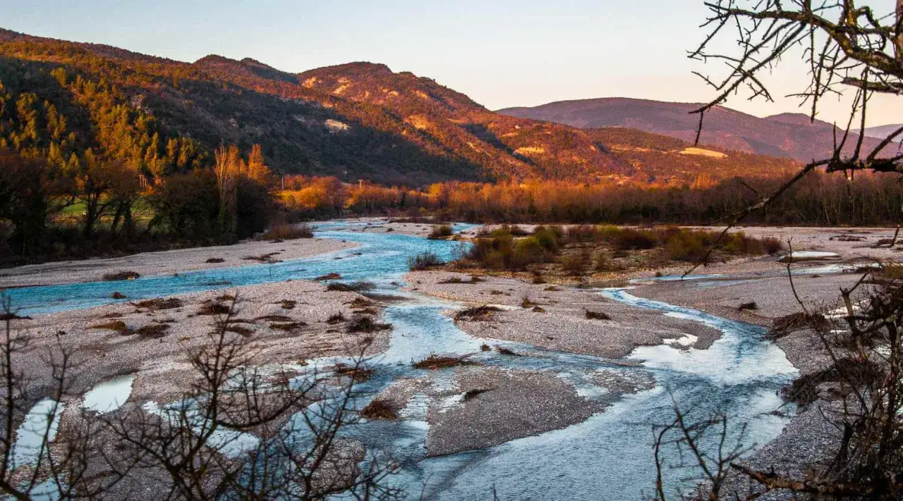 Le saut de la Drôme, un paysage de rivière aux eaux bleues vives serpentant entre des bancs de galets, sous des montagnes dorées par le soleil couchant.