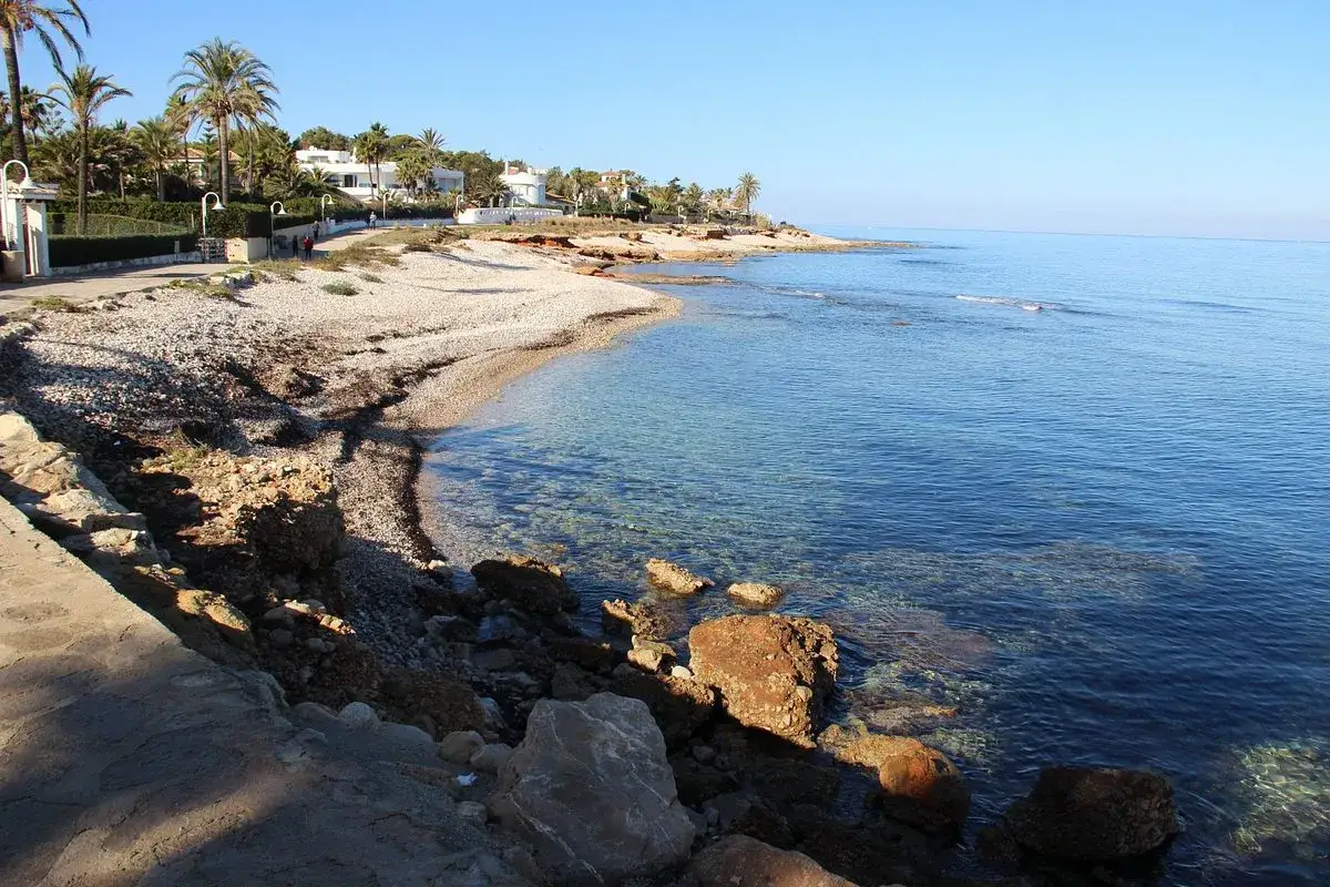 Costa rocosa con playa de guijarros y aguas cristalinas. Palmeras y casas blancas se ven al fondo, cerca de la **punta negra**.