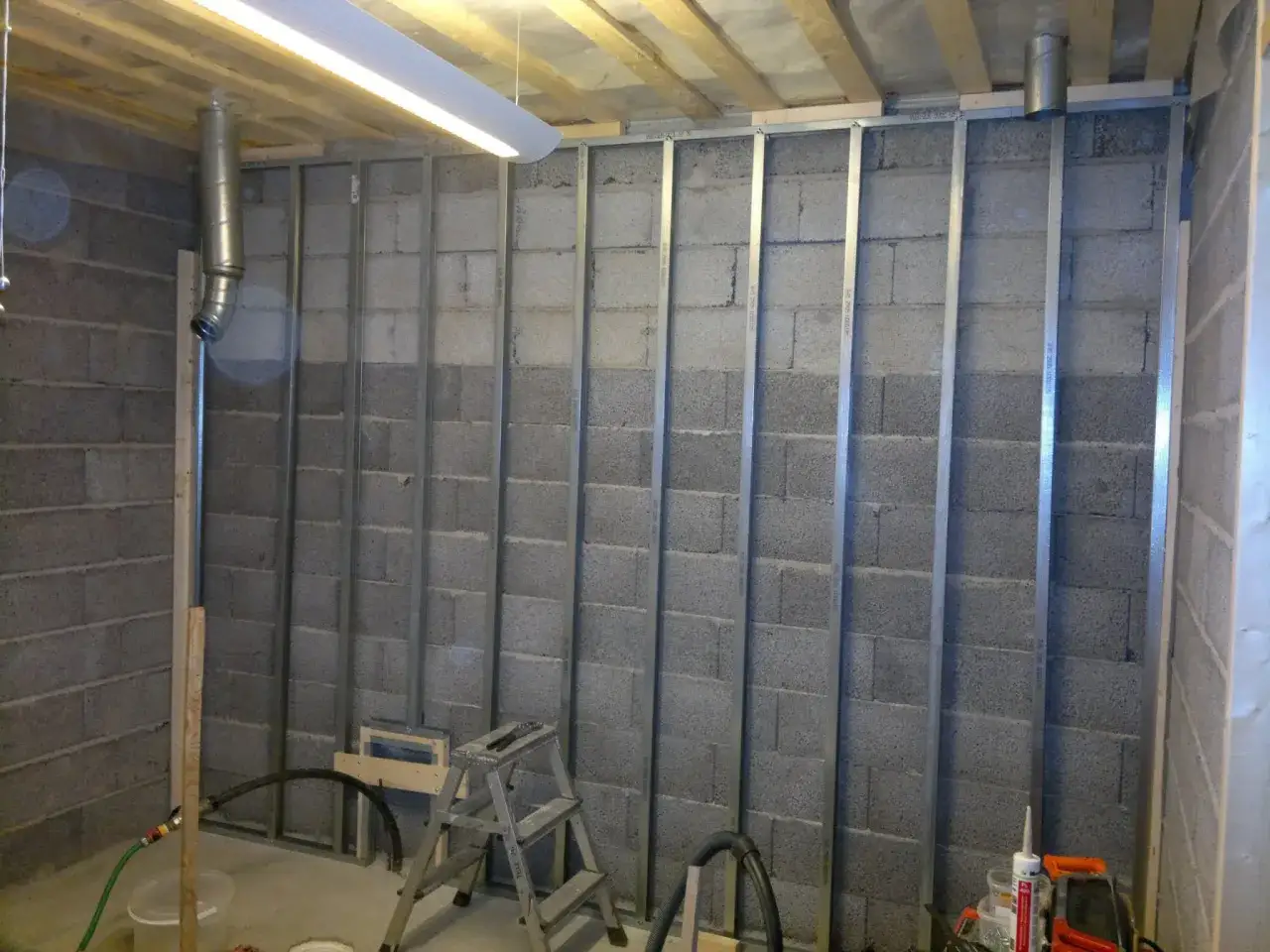 Arbetet med att montera stålreglar pågår mot en betongvägg. En stege och verktyg syns i förgrunden.