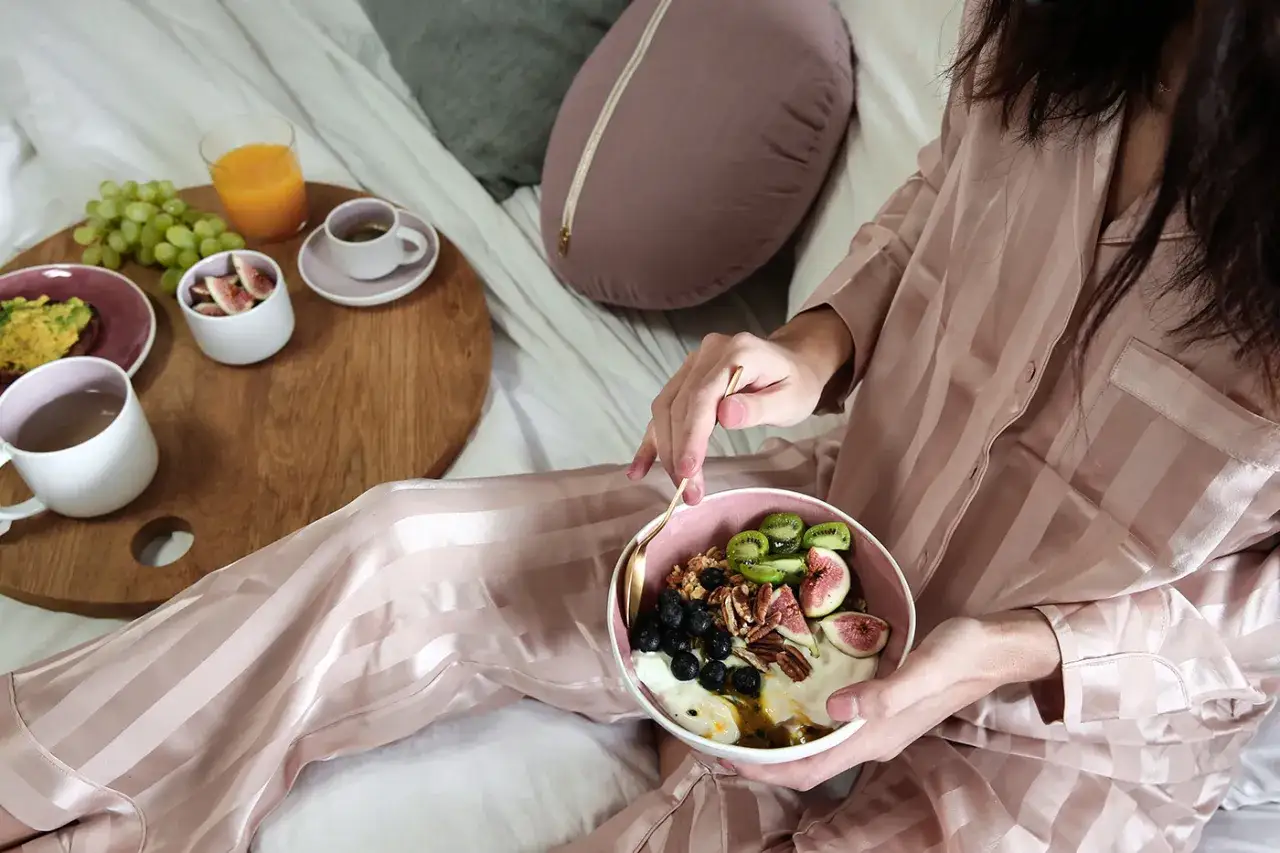 A person in silky pajamas enjoys a healthy breakfast of yogurt, fruit, and granola, a moment to seek contentment.