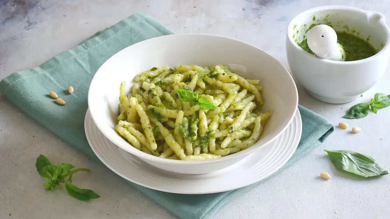 Un piatto invitante di pasta al pesto genovese, con basilico fresco e pinoli sparsi. Accanto, un mortaio con il pesto appena fatto.