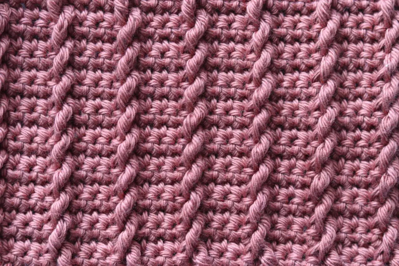Detaljerad bild av virkat tyg i en rosa nyans. M&ouml;nstret best&aring;r av diagonala fl&auml;tor skapade med tvinnat garn, som ger en texturrik yta.