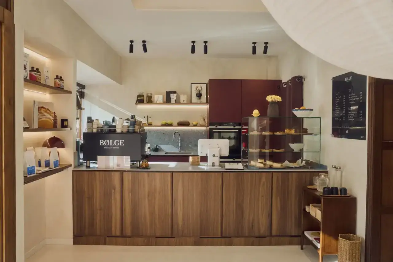 Interior de una cafeter&iacute;a moderna, ideal para quienes buscan c&oacute;mo abrir una cafeter&iacute;a en Espa&ntilde;a. M&aacute;quina de caf&eacute;, vitrina de pasteles y estanter&iacute;as.