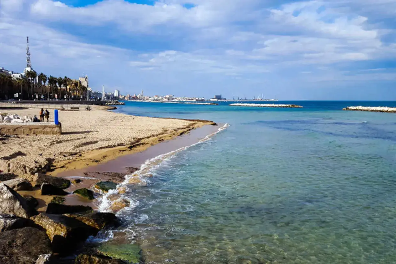 Vue sur la bari plage avec des vagues douces, des rochers et la ville en arrière-plan sous un ciel bleu et nuageux.
