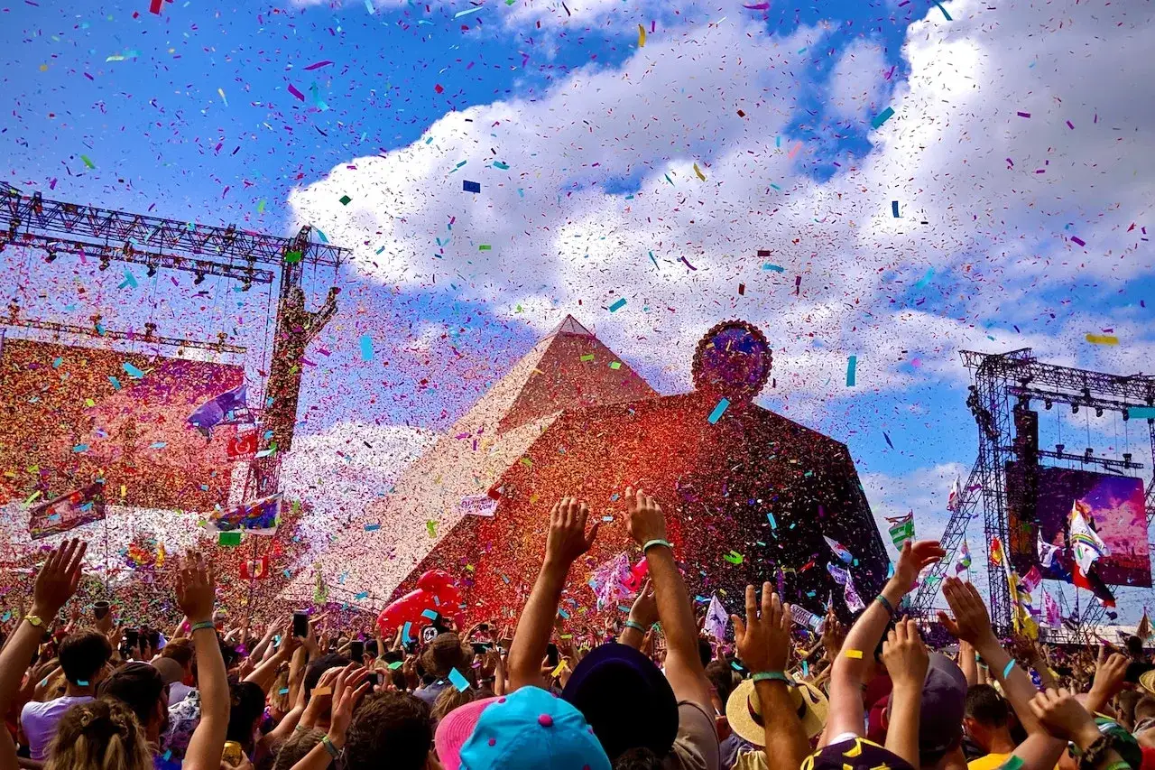 Confetti rains down on a massive crowd at a large festival in the United Kingdom, with the iconic Pyramid stage in the background.