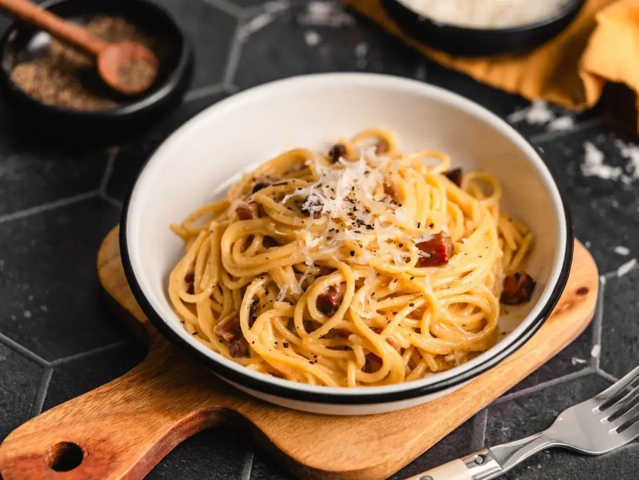 Eine Sch&uuml;ssel mit echter Carbonara, garniert mit geriebenem Parmesan und schwarzem Pfeffer, serviert auf einem Holzbrett.