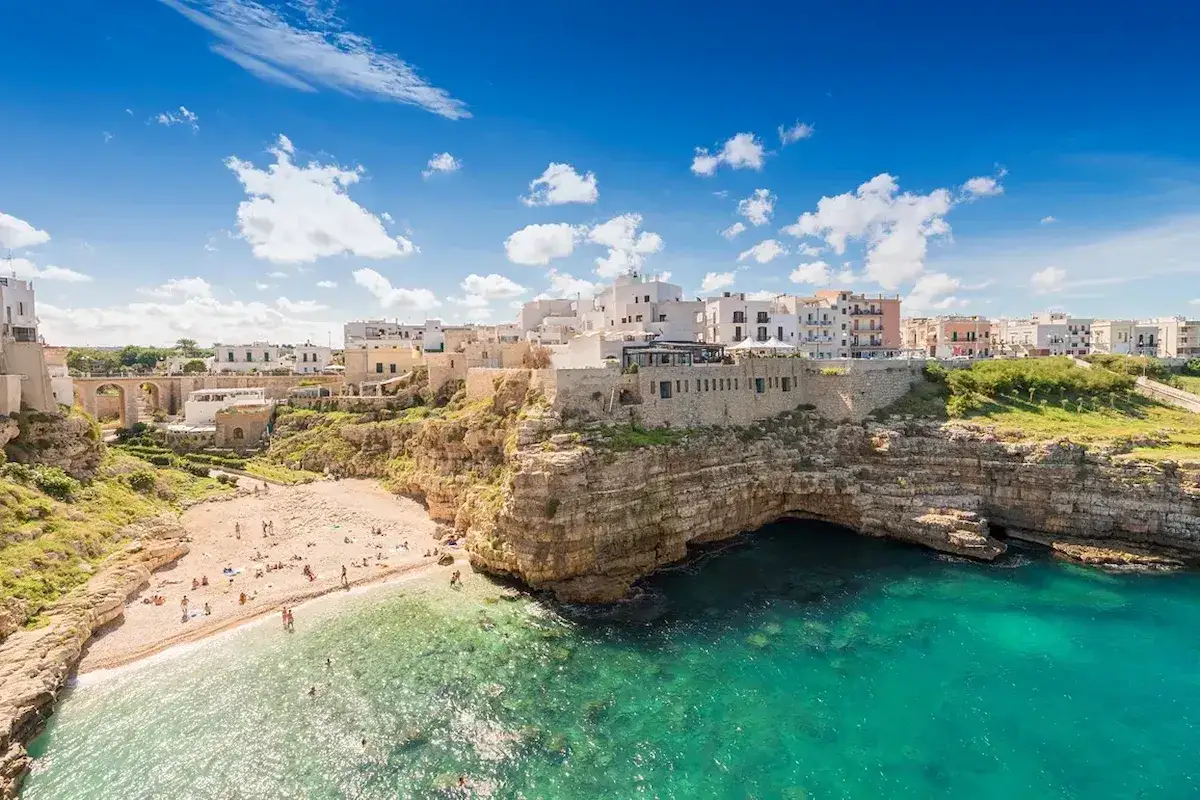Vackra vita hus kl&auml;ttrar uppf&ouml;r klipporna vid Polignano a Mare, med en solig strand och turkost vatten.