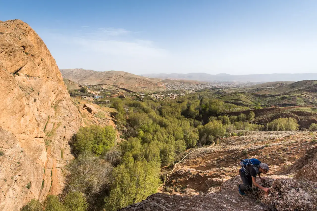 Wanderer erklimmt felsigen Hang im **Gebirge im Iran** mit Blick auf ein gr&uuml;nes Tal und eine Stadt.