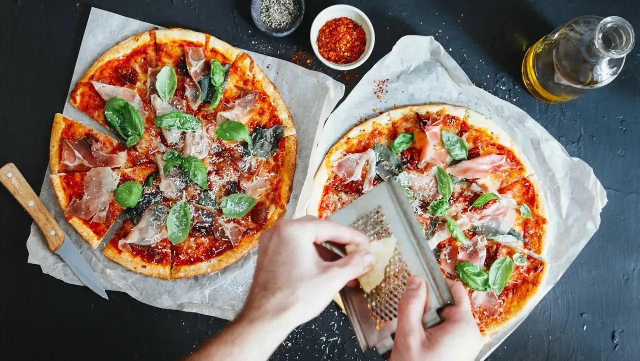 Dwie pyszne pizze z szynką parmeńską i bazylią, posypywane serem. W tle przyprawy i oliwa, idealne dodatki do pizzy.