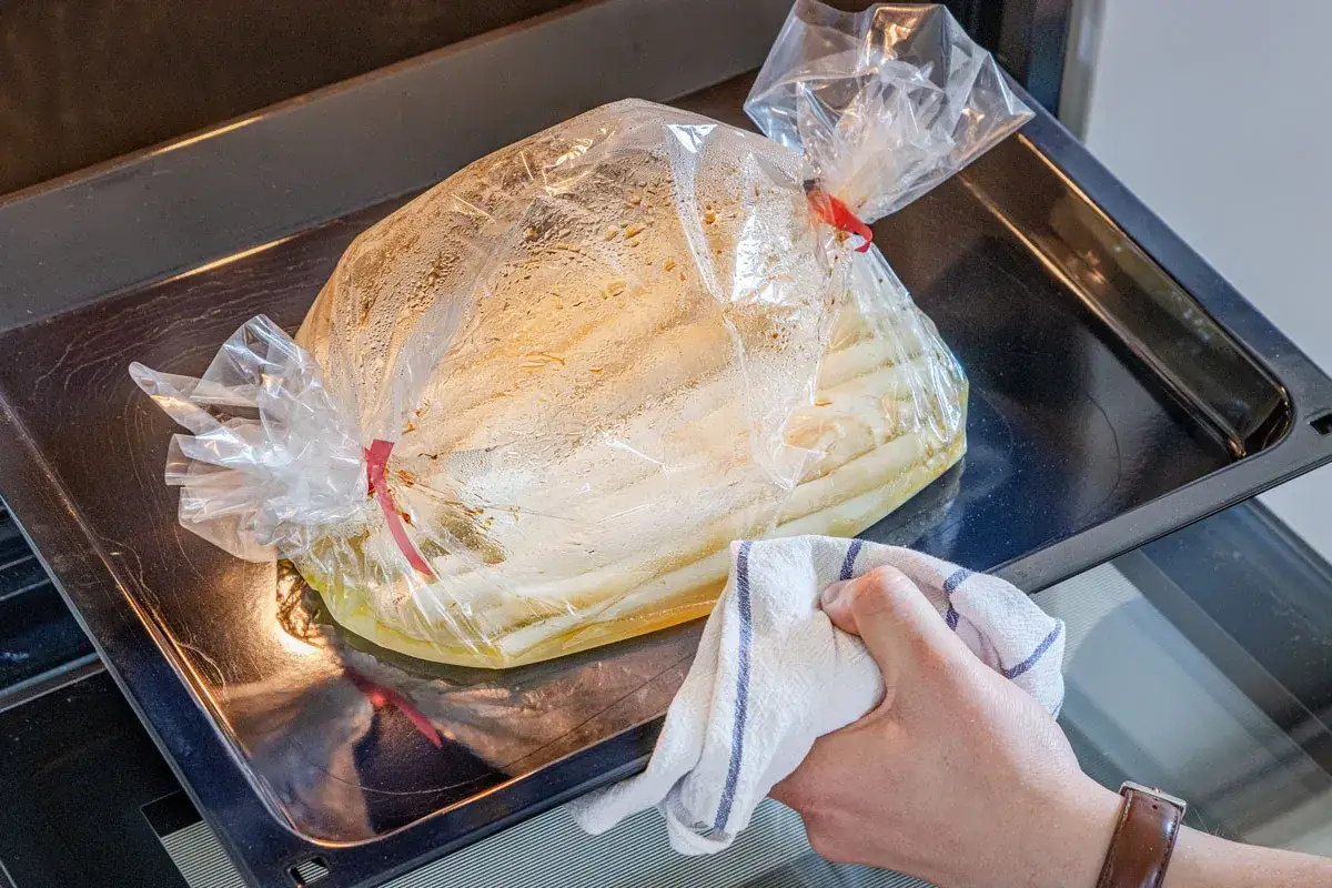 Spargel im Bratschlauch dampft auf einem Backblech. Eine Hand mit K&uuml;chentuch schiebt das Blech in den Ofen.