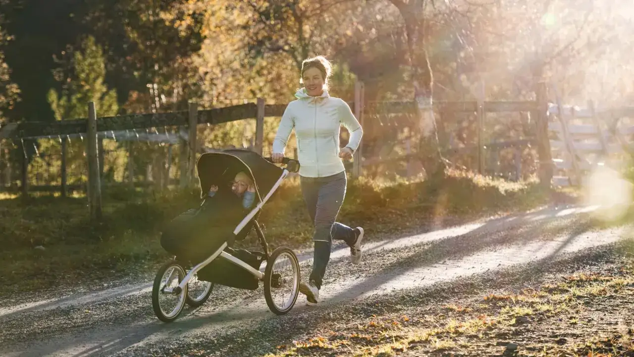 Frau joggt mit Kinderwagen auf einem Feldweg im goldenen Licht der Herbstsonne.