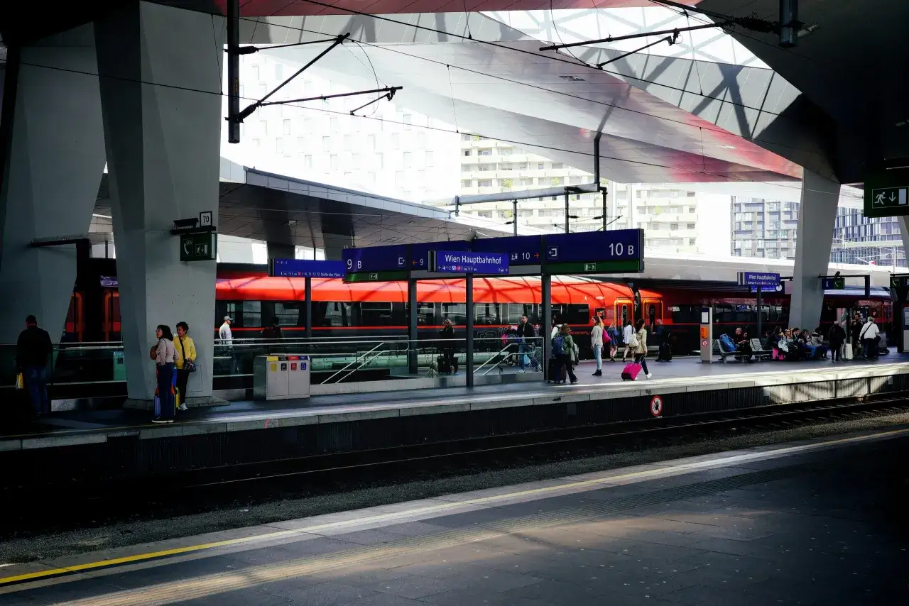 Menschen warten auf dem Bahnsteig f&uuml;r ihre zugreise nach wien. Ein roter Zug steht bereit.
