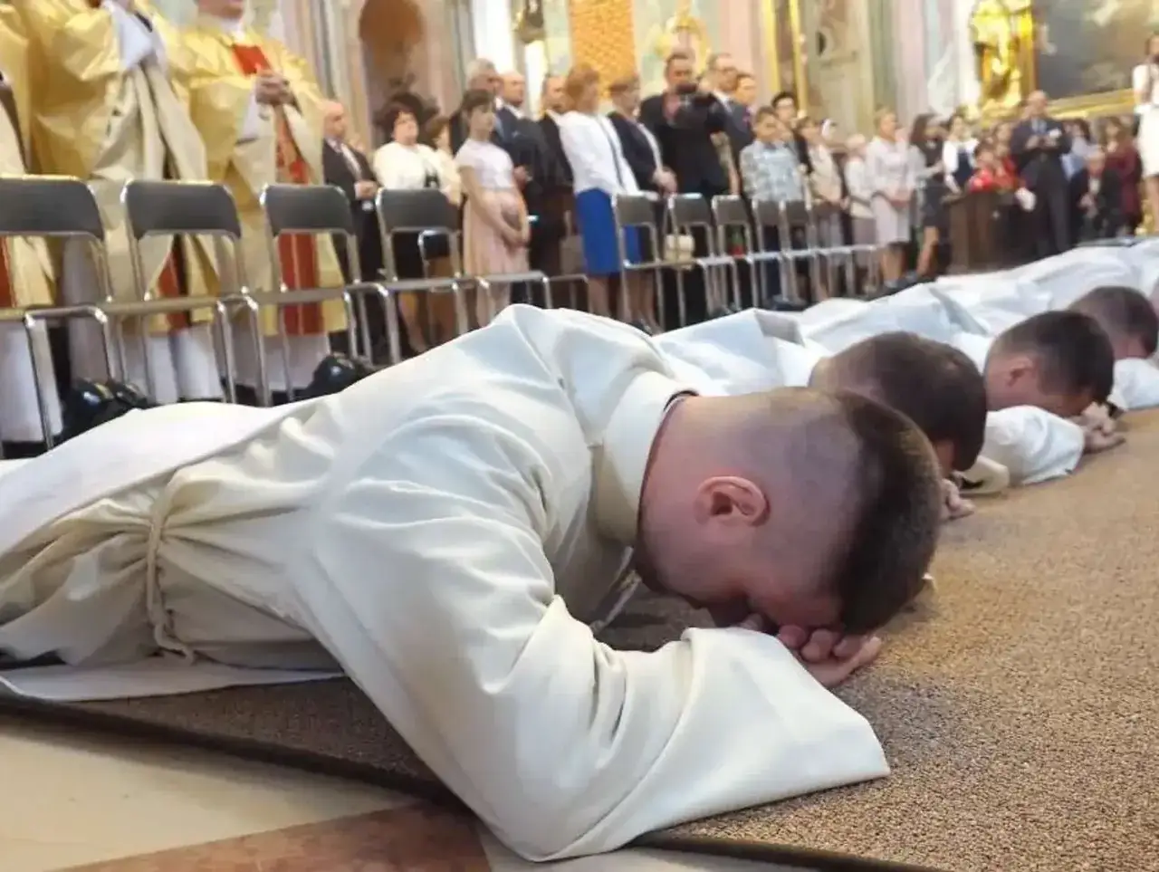 Klerycy w białych szatach klęczą na dywanie. W tle zgromadzeni wierni. Czy wdowiec może zostać księdzem? Tak, w pewnych okolicznościach.
