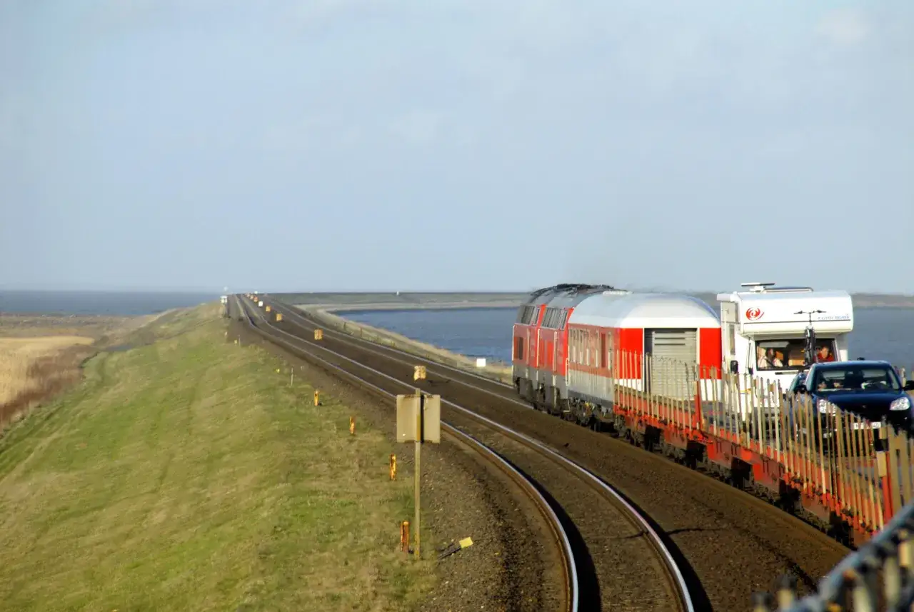 Ein roter Zug fährt auf einem Bahndamm mit Blick auf das Meer. Autos und ein Wohnmobil sind auf einem Waggon geladen. Eine Reise mit dem Zug durch Deutschland.