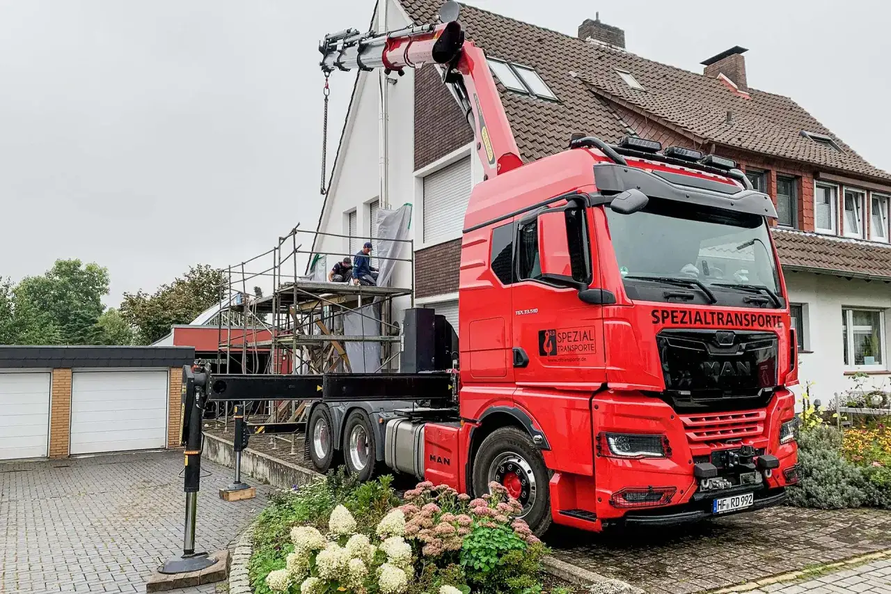 Ein roter LKW mit einem Kranarm, der auf einem Ger&uuml;st &uuml;ber einem Haus schwebt. Das zeigt, was ist ein Kran: ein m&auml;chtiges Werkzeug f&uuml;r schwere Hebearbeiten.