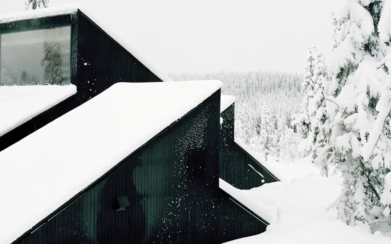 Une cabane semi enterrée aux lignes modernes, recouverte de neige, se dresse dans un paysage forestier hivernal.