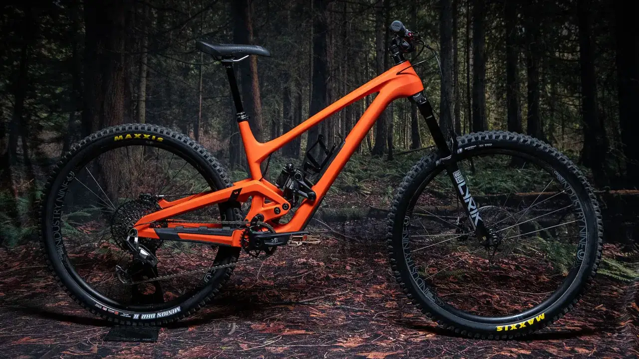 An evil offering review: a bright orange full-suspension mountain bike with Maxxis tires and Industry Nine wheels, set against a dark forest backdrop.