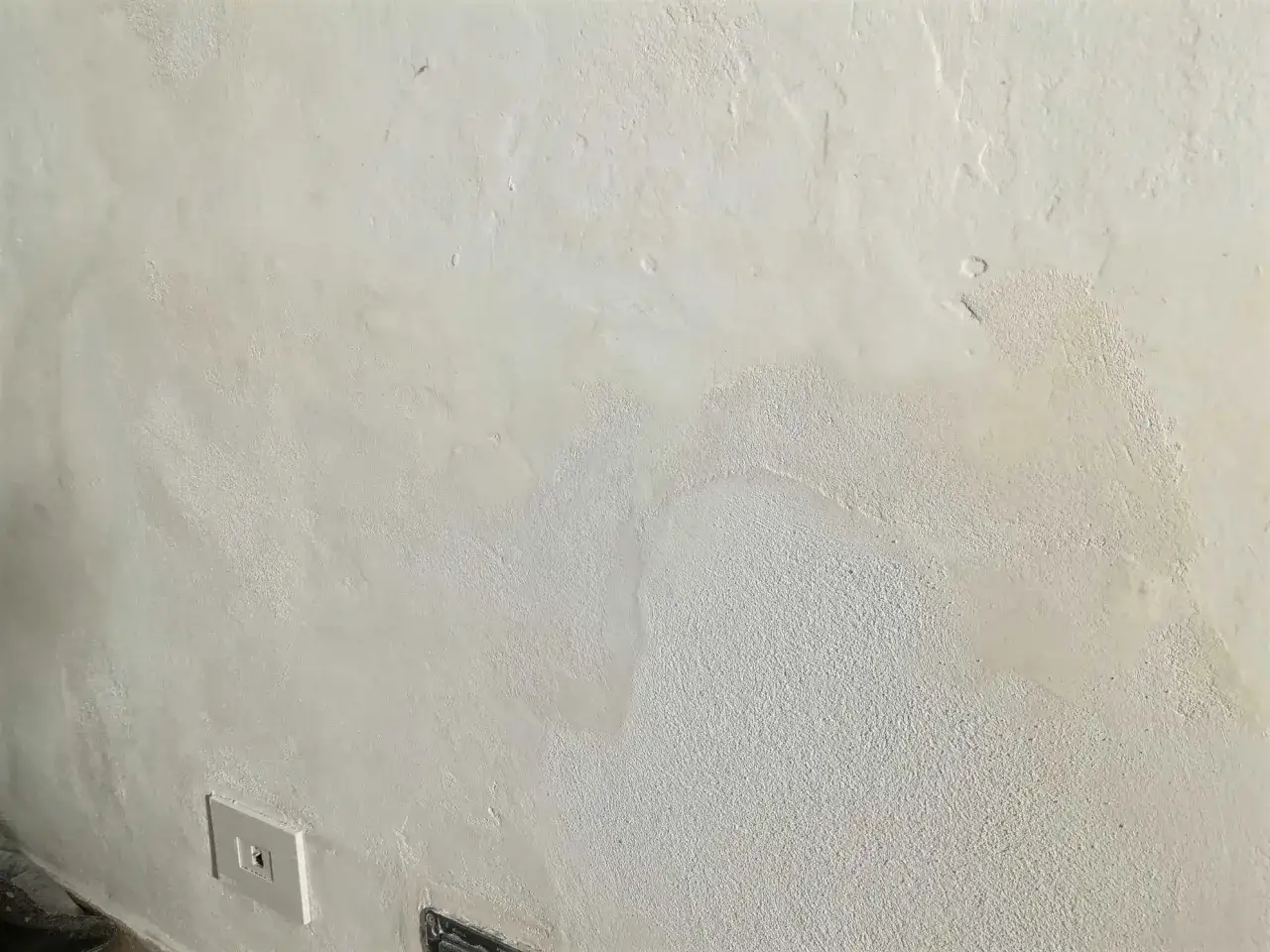 Muro in fase di rasatura, con diverse texture e una presa elettrica visibile.
