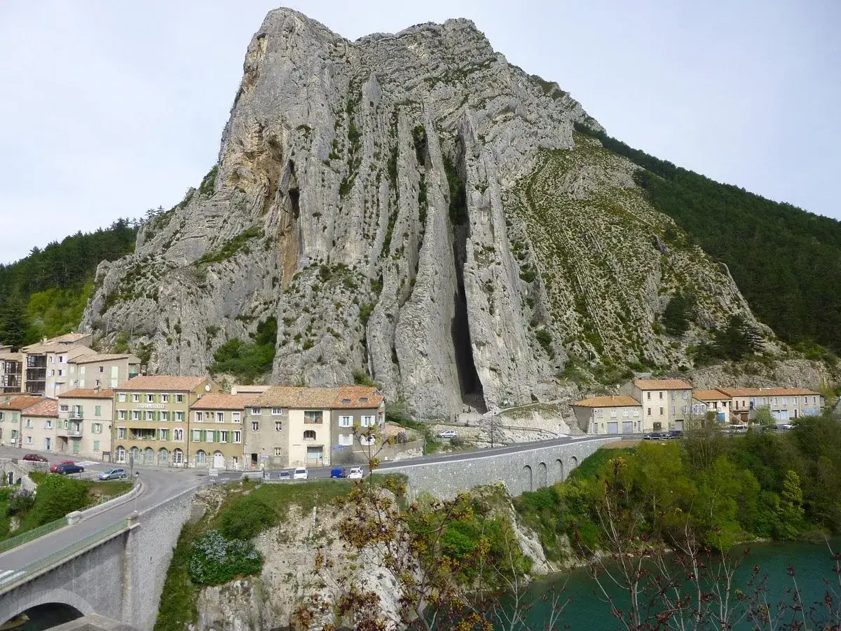 Imponerande klippformation vid Route Napol&eacute;on. En by kl&auml;ttrar uppf&ouml;r bergssluttningen, med en bro och en turkos flod nedanf&ouml;r.