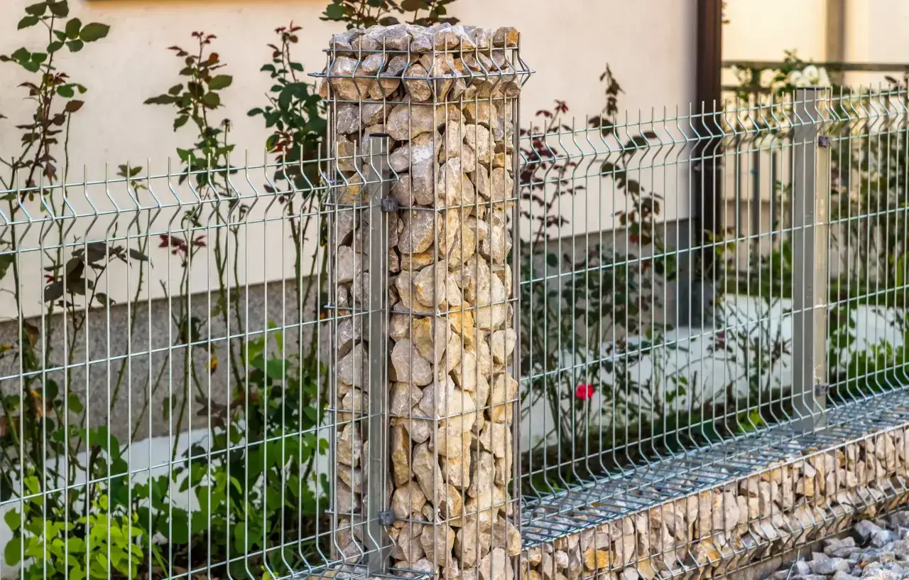Moderne Ideen f&uuml;r eine Gartenmauer: Ein Gabionenpfeiler aus Natursteinen neben einem Drahtgitterzaun, umgeben von Rosenb&uuml;schen.
