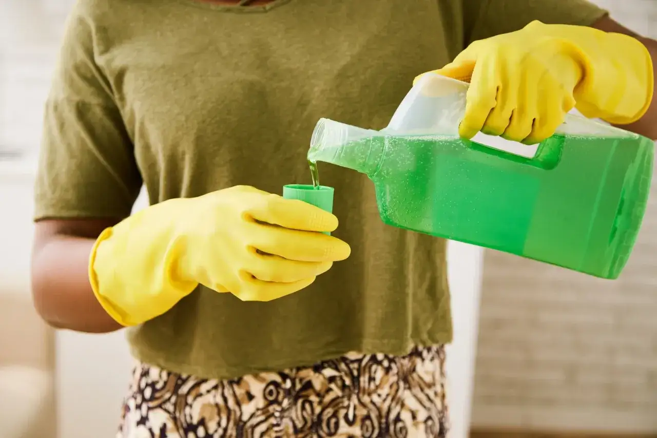 Ręce w żółtych rękawiczkach nalewają zielony płyn z dużego pojemnika do mniejszej miarki. To może być sposób, jak usunąć kamień z plastiku.