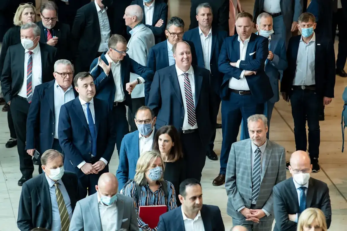 Abgeordnete im Bundestag, einige mit Masken, stehen dicht gedrängt. Ein Mann in dunklem Anzug und gestreiftem Hemd blickt nach vorne. Der **Bundestag Hammelsprung** ist ein wichtiger Moment der Abstimmung.