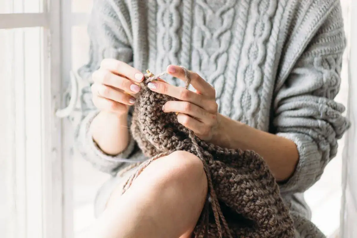 Mani che lavorano a maglia con filato marrone, indossando un maglione grigio a trecce. Un momento di tranquillità per lavare lana a mano.