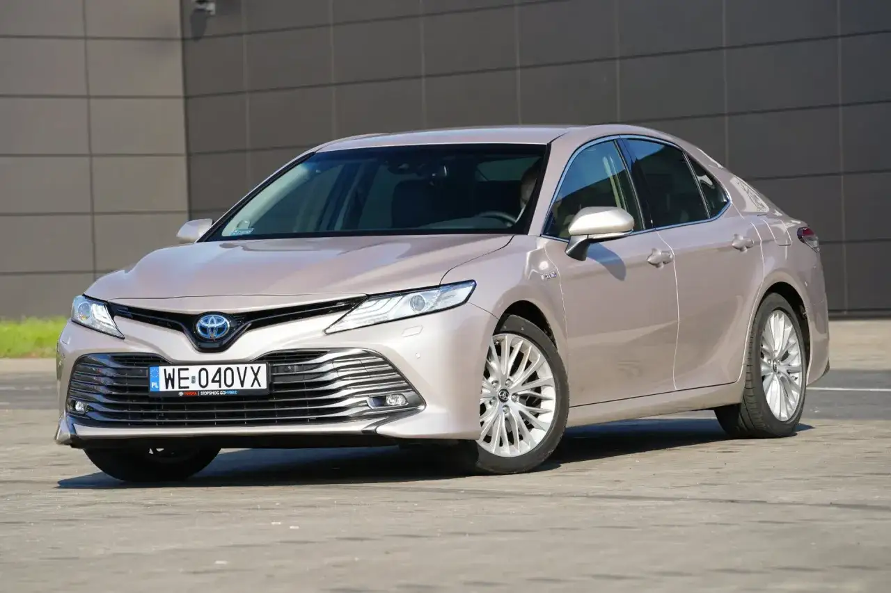 Elegancka, beżowa Toyota Camry Hybrid zaparkowana przed nowoczesnym budynkiem. Poznaj jej imponujące toyota camry wymiary.