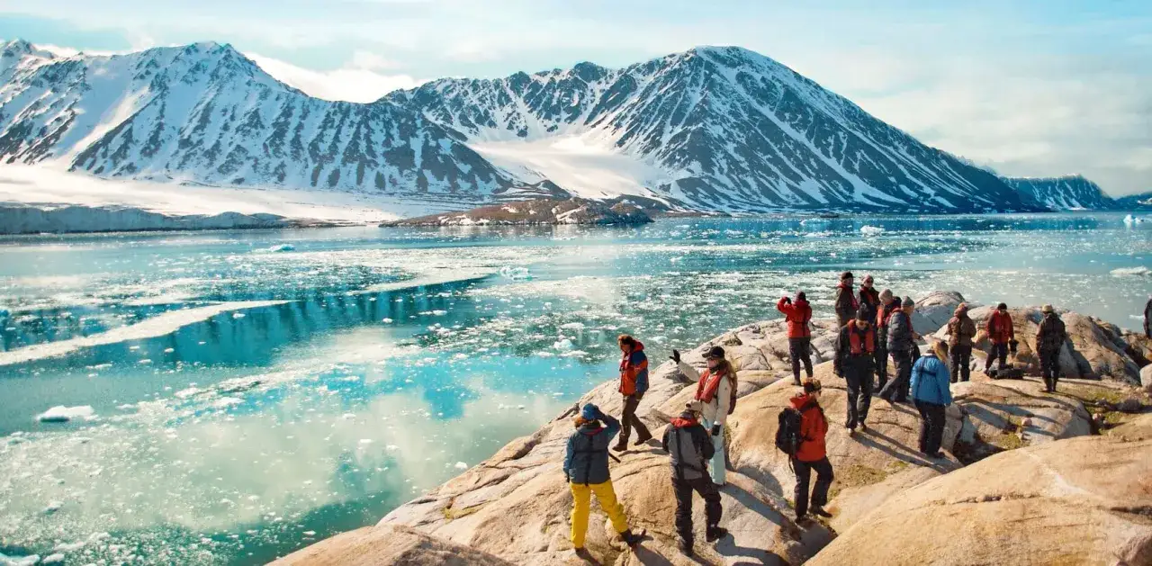 Grupa turystów na skalistym brzegu obserwuje lodowate wody i ośnieżone góry. Spitsbergen wycieczka cena - niezapomniane widoki.