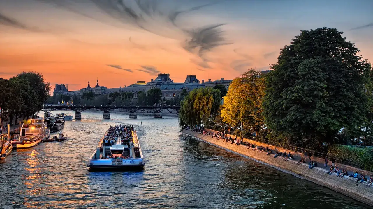 Coucher de soleil sur la Seine, un bateau plein de touristes navigue près d'un bar sur les quais parisiens. Des gens profitent de la douceur du soir.