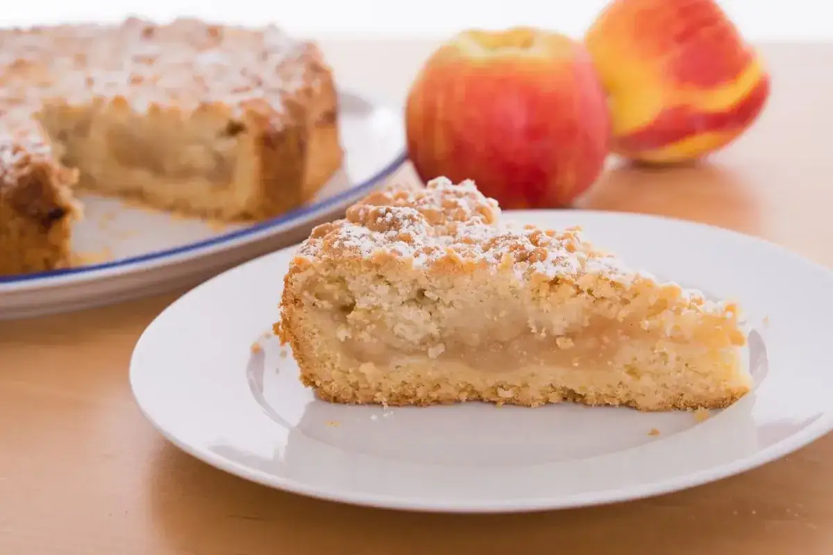 Una fetta di crumble di mele, cosparsa di zucchero a velo, su un piatto bianco. Sullo sfondo, una torta intera e due mele rosse.