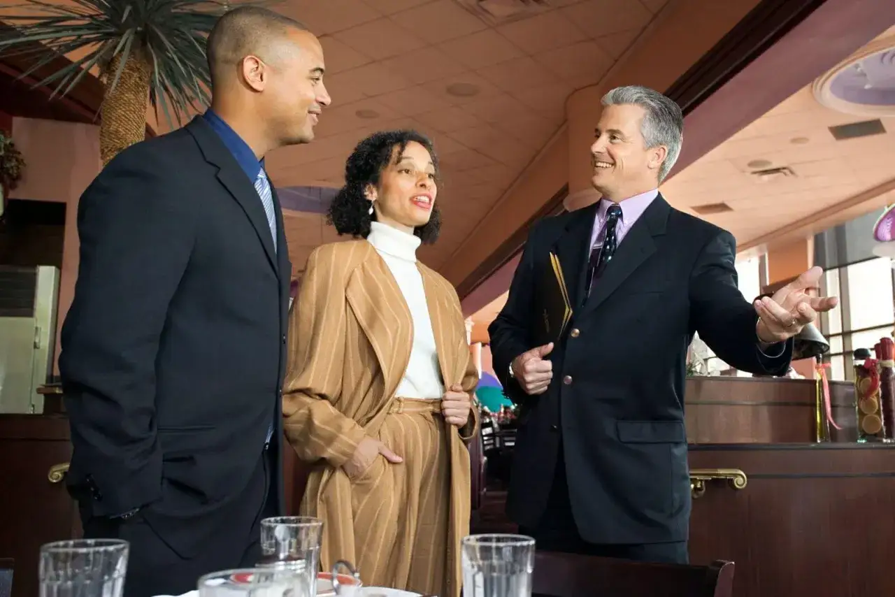 Tres personas conversan en un restaurante. Uno de ellos, con un traje marrón a rayas, parece explicar algo, quizás sobre el menú o el **metre de cocina**.