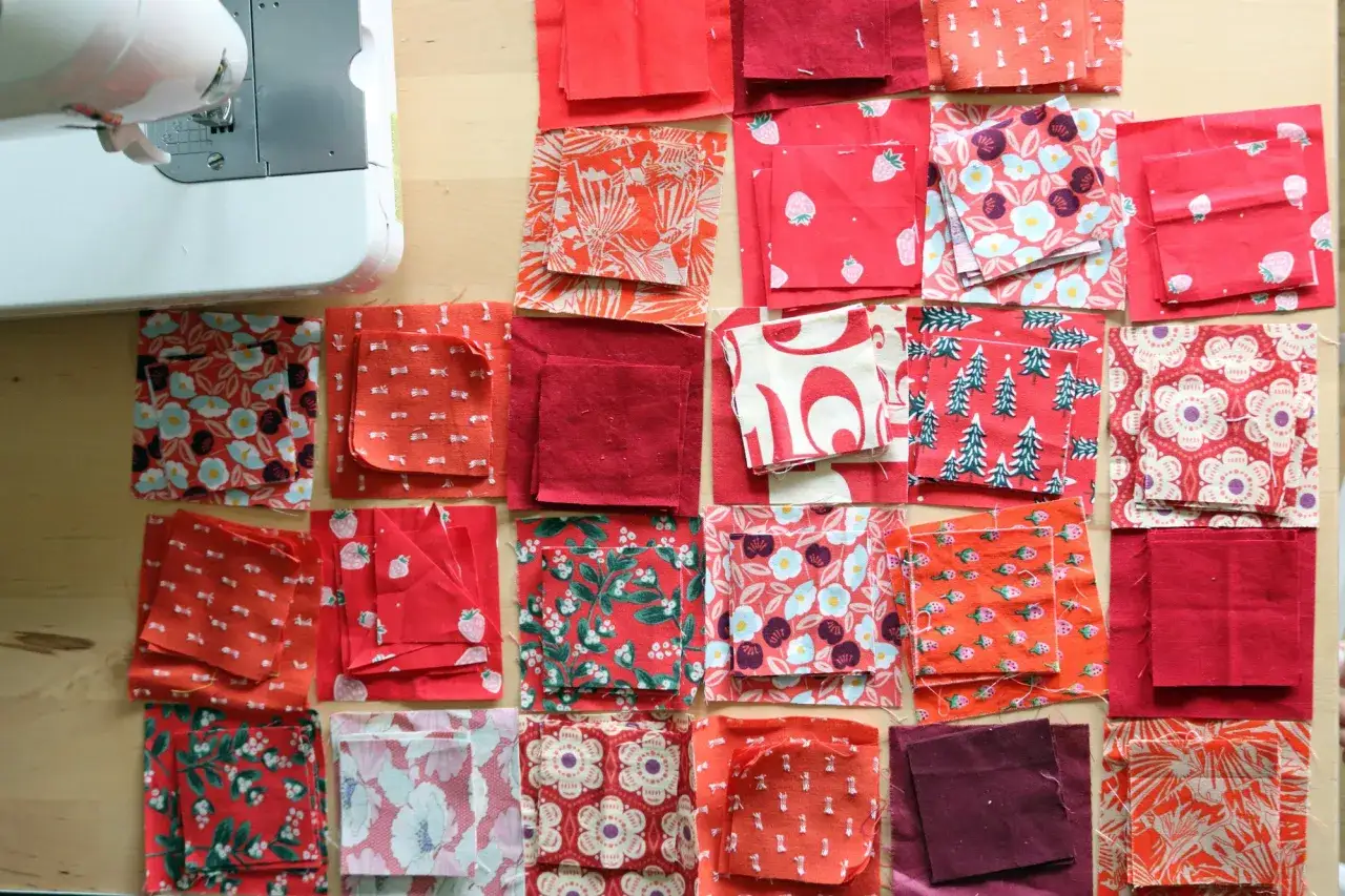 Viele rote Stoffquadrate mit Mustern wie Blumen, Erdbeeren und B&auml;umen liegen bereit f&uuml;r neue **patchwork ideen**. Eine N&auml;hmaschine ist im Hintergrund zu sehen.