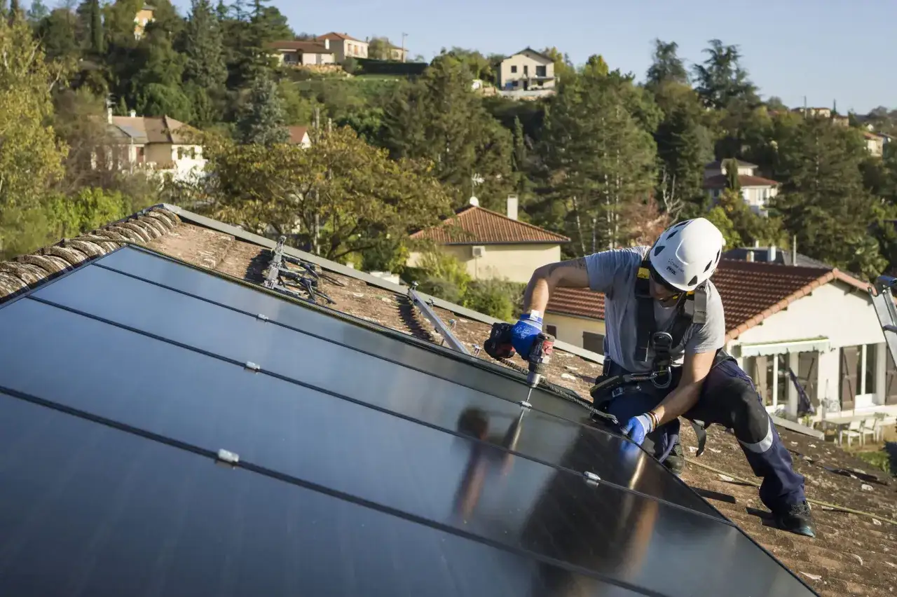 Un homme en casque et harnais installe des panneaux solaires sur un toit. Il utilise une perceuse pour fixer les éléments, une étape clé pour apprendre comment installer des panneaux solaires soi même.