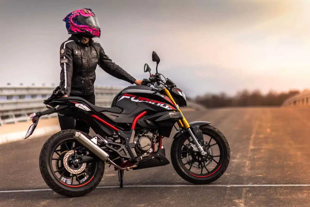 Motocyklistka w różowym kasku i czarnej kurtce opiera się o sportowy motocykl. Kategoria AM.
