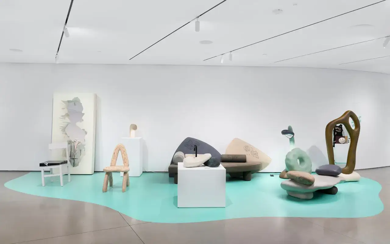 Ausstellung mit surrealen M&ouml;beln und Skulpturen, die wie organische Formen wirken. Ein Spiegel mit Silhouette, ein Holzstuhl und eine gr&uuml;ne, donut-&auml;hnliche Skulptur sind Teil dieser aktuellen Kunst.